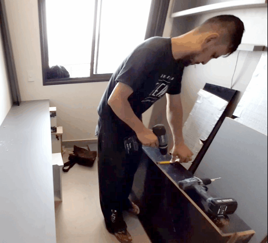 Montador de móveis medindo e furando painel de madeira com parafusadeira em quarto com janela, durante instalação de marcenaria planejada